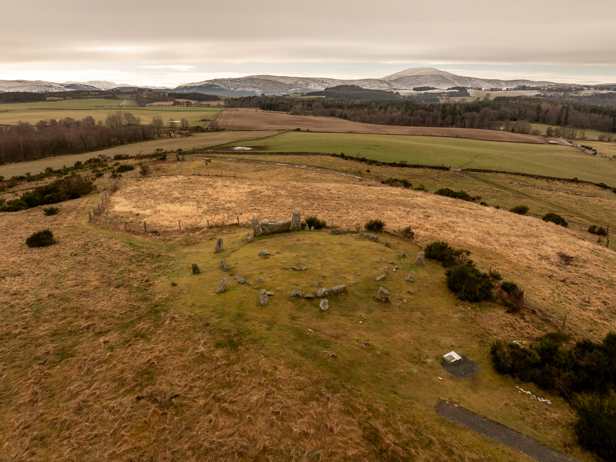 A drone photo of a stone circle on a small exposed hill. In the background there is a snow-capped mountain. 
