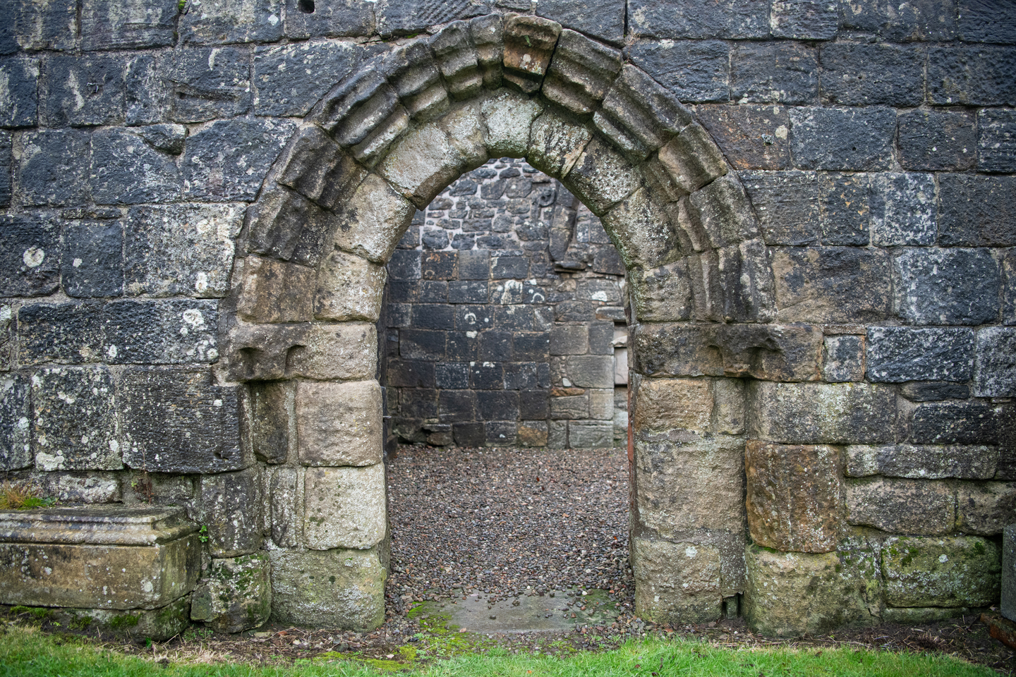 A medieval stone archway in an old stone wall, leading to a gravel floor inside the structure. The stones are worn and uneven, showing significant age.