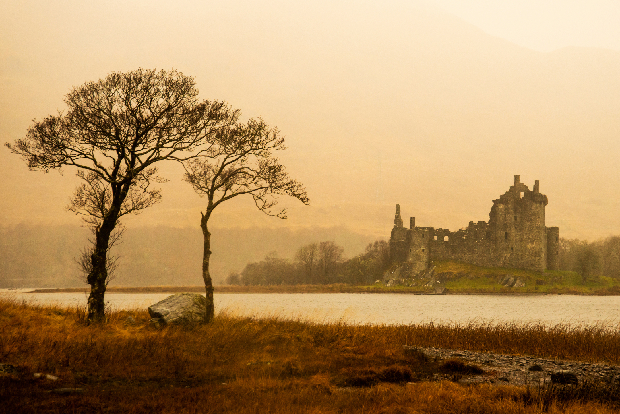 Spooky, dark, orange-toned view of castle across a loch, with trees bare of leaves