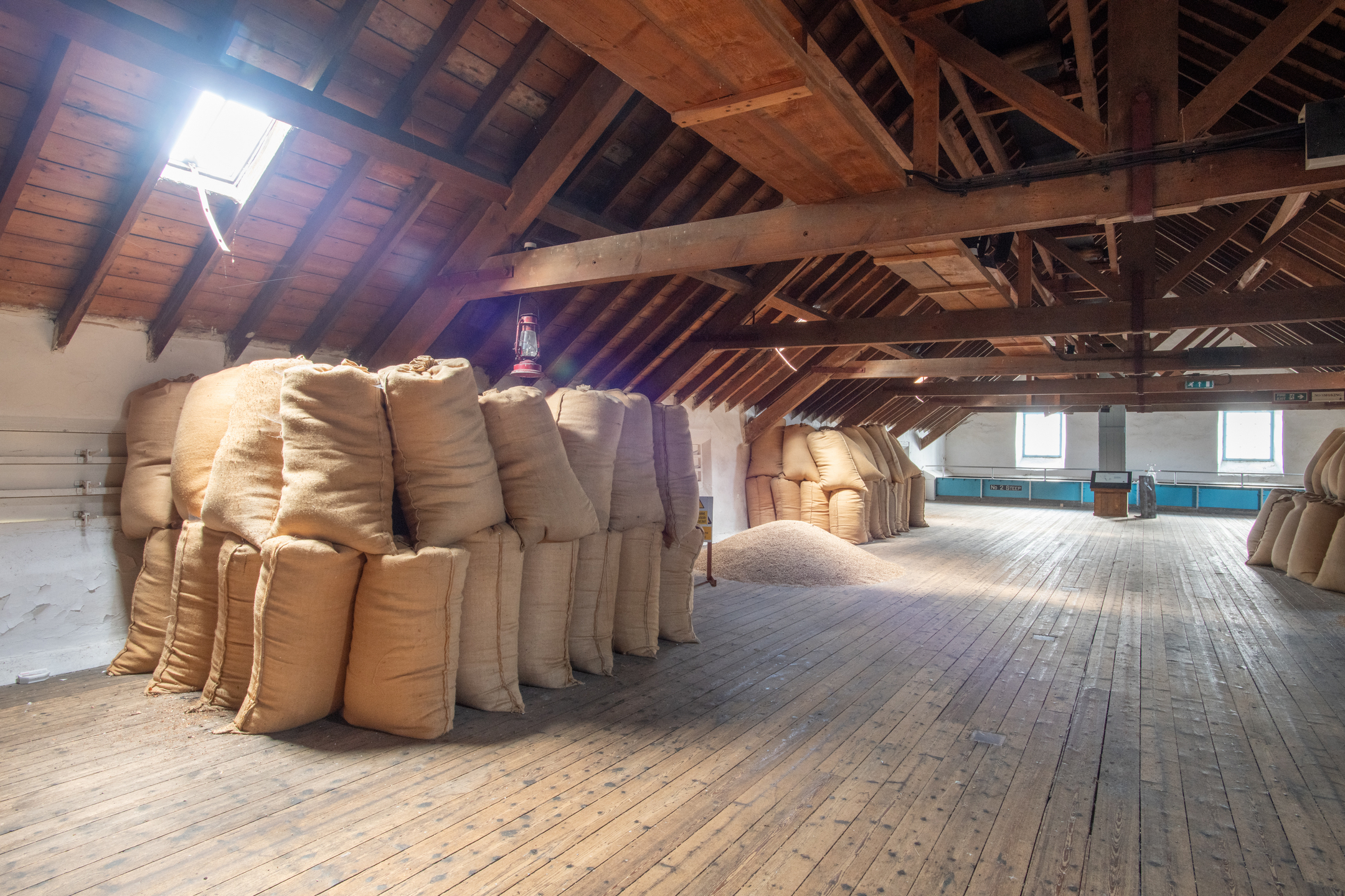 Interior of a traditional malt loft at Dallas Dhu distillery, with stacks of grain sacks arranged beneath exposed timber roof beams and skylights.