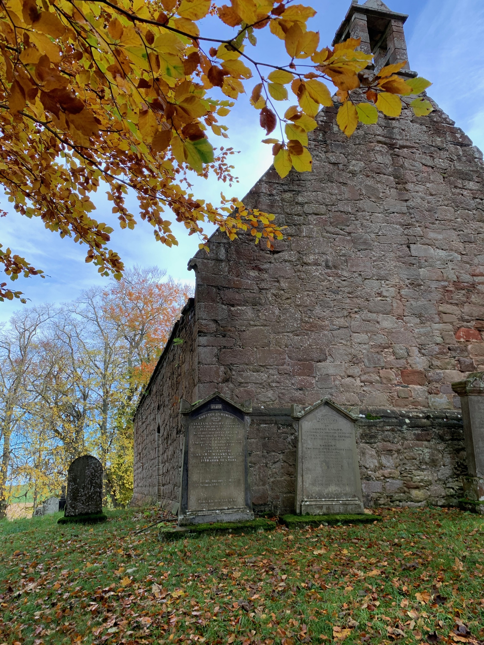 gable end of a stone church with bell tower and grave stones, and autumn leaves filling the top left corner of the frame