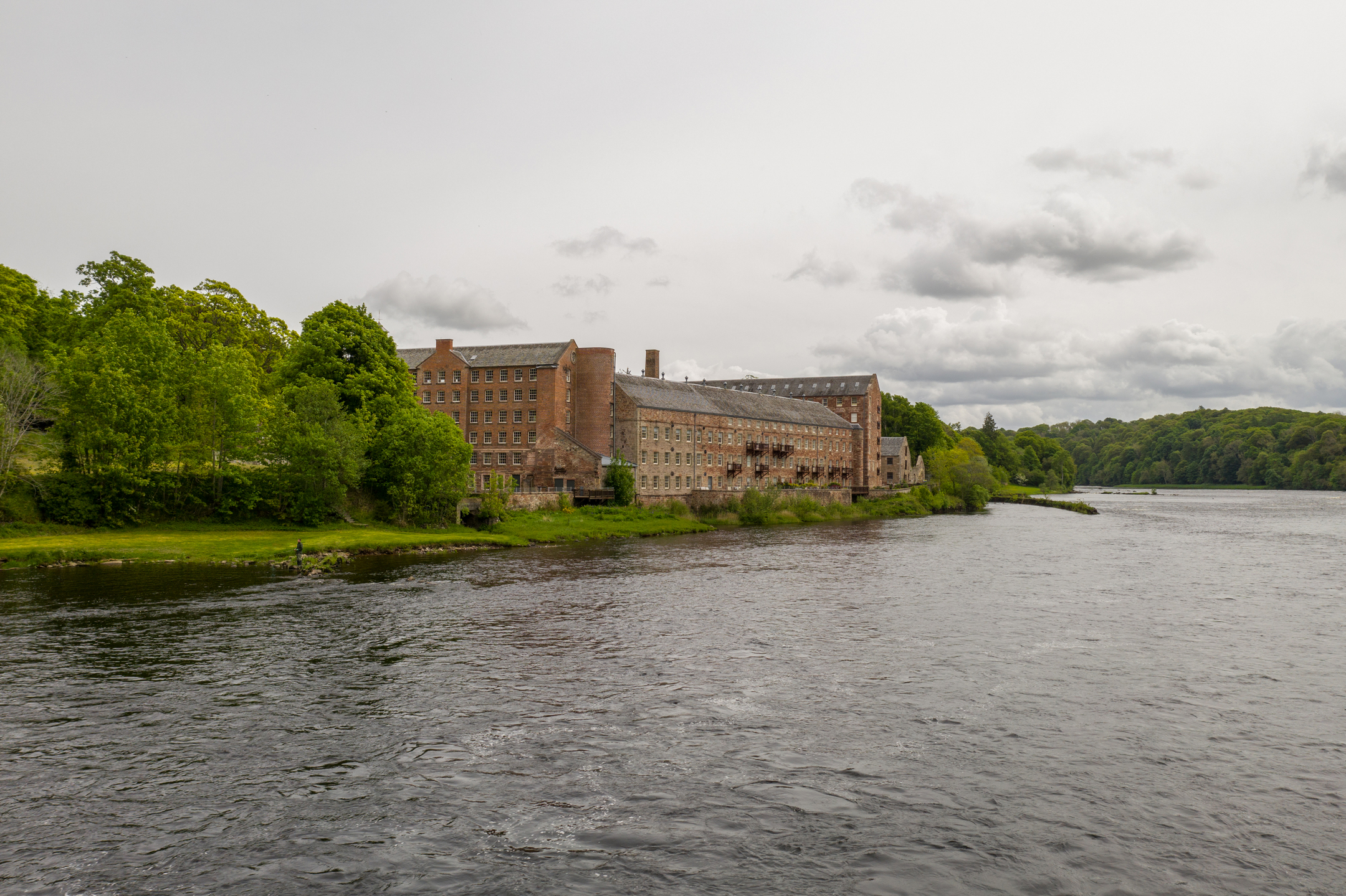 A riverbank view of the long brick Stanley Mills buildings surrounded by trees, with the river flowing beside them under an overcast sky.