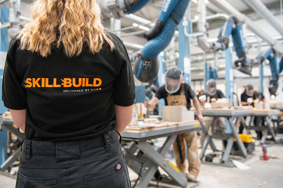 A person with long blond hair stands with back to photographer weather a black t-shirt with 'skill build' written on the back. In front of them, apprentice stonemasons work while stood at rows of tables.