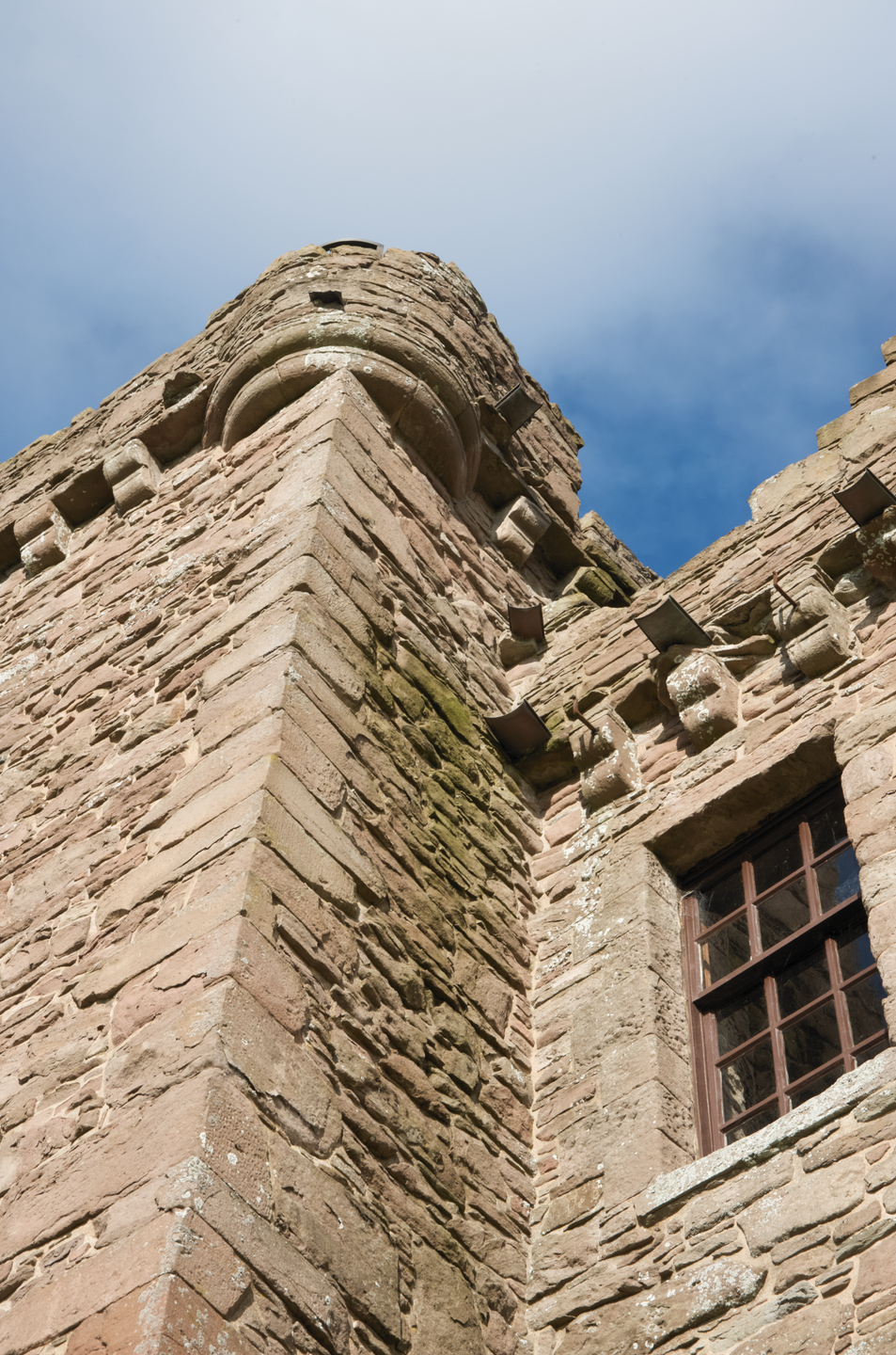 Looking up at the side of a stone building. There is some moss lightly growing on the stone. There is a window with wood panelling. It suggests an old building. 