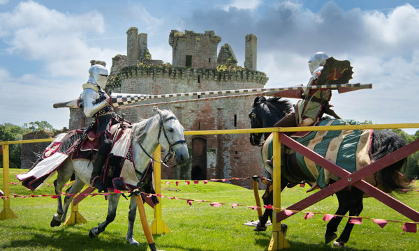 Two knights on horses racing towards each other with their lances raised and a castle in the background on a bright sunny day