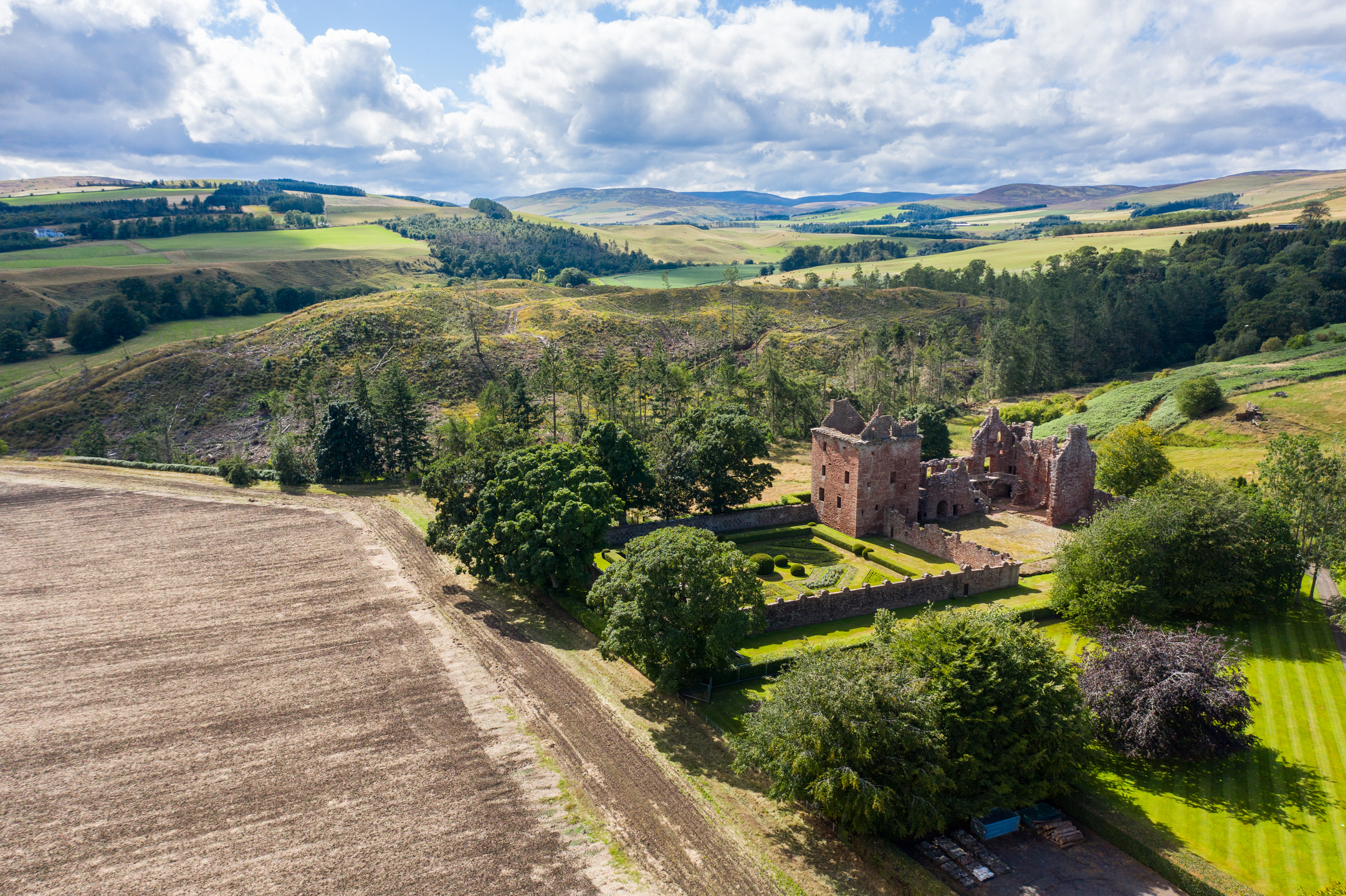 An aerial photo of a ruined castle and a walled garden nestled amongst trees. It is surrounded by picturesque farmland and rolling hills. 