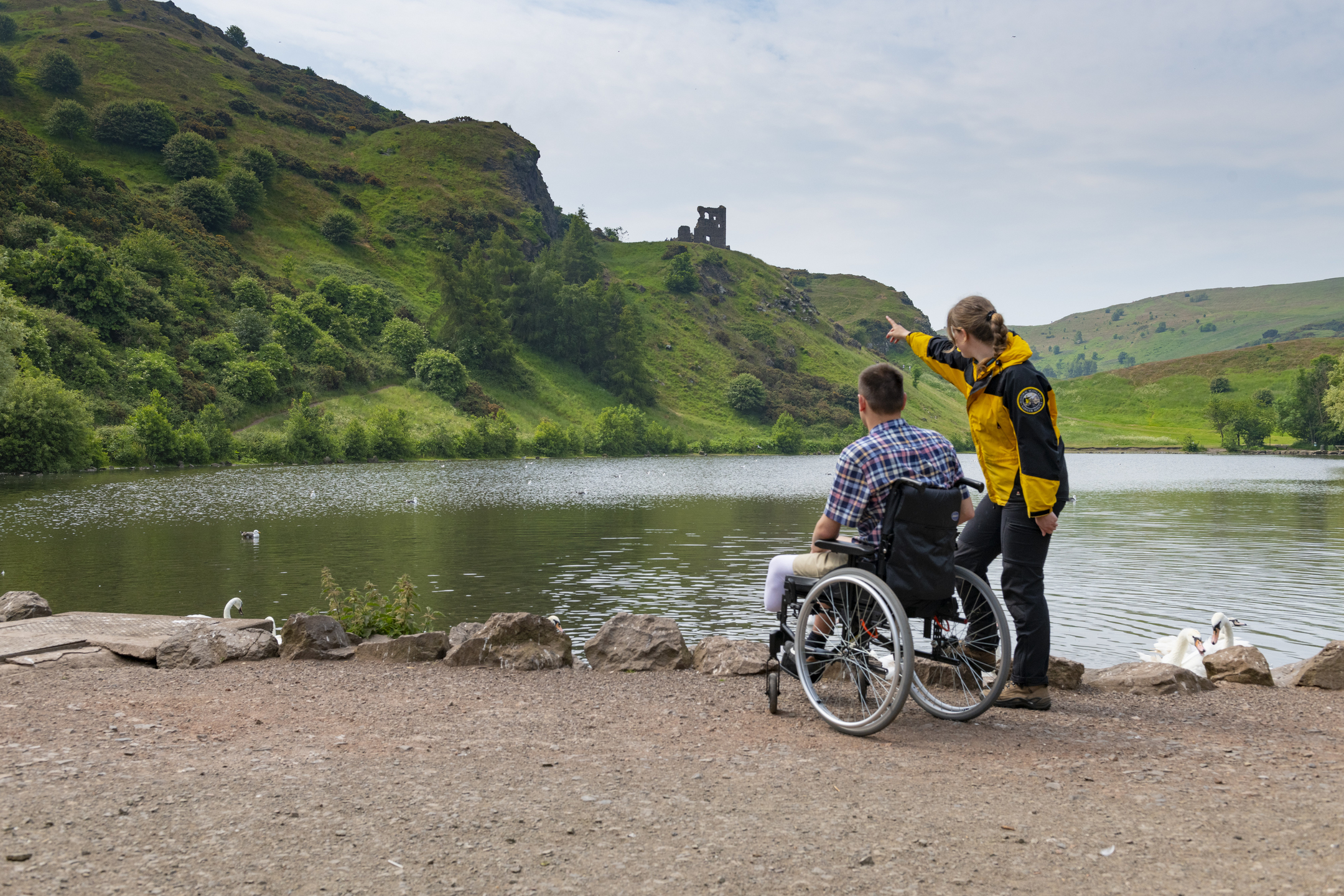 Assistant ranger and visitor in a wheelchair by a lake. The ranger is pointing to a ruin high on a hill in the distance. 