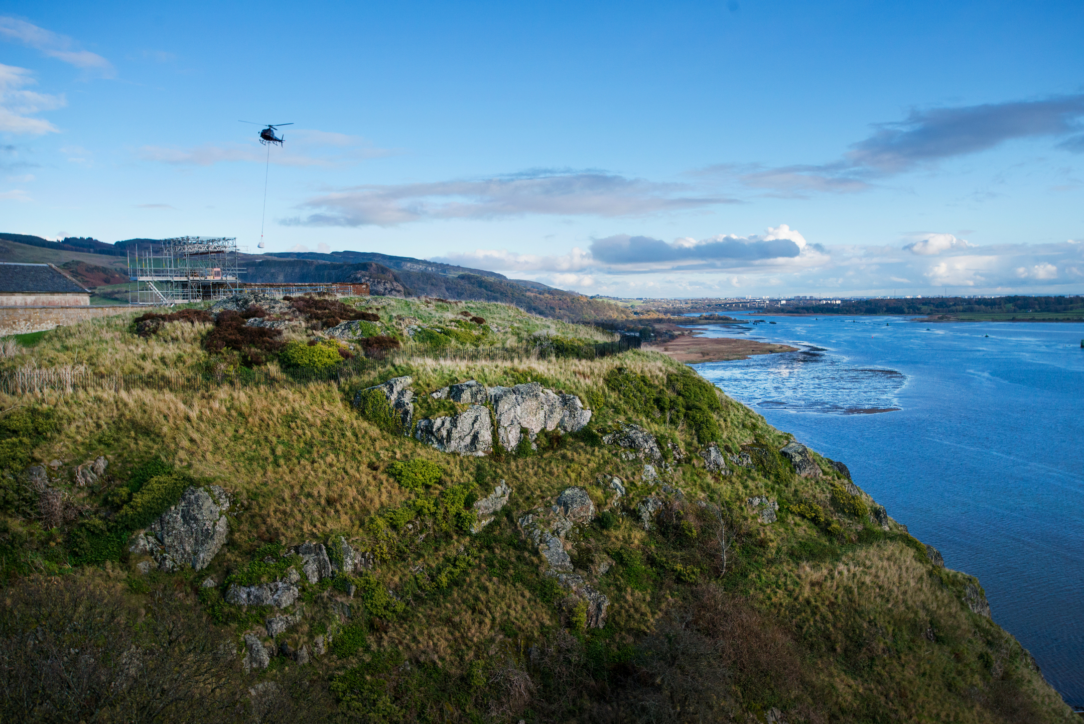A helicopter carries cargo beneath it while flying over grassy, rugged terrain near the river and castle.