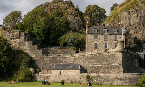 A stone castle with grass in the front and people sitting on picnic benches and walking on the grass. On either side of a stone house there is a large rock face covered with vegetation. In front of the house there is a saltire flag flying.