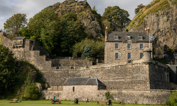 A stone castle with grass in the front and people sitting on picnic benches and walking on the grass. On either side of a stone house there is a large rock face covered with vegetation. In front of the house there is a saltire flag flying.