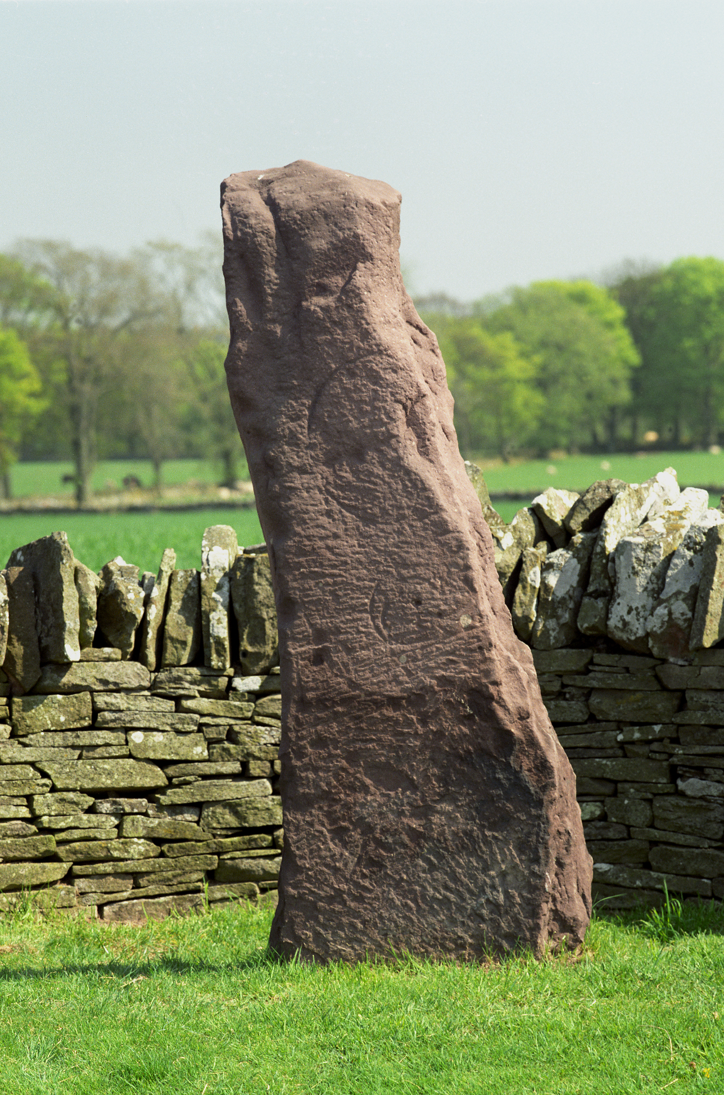 An ancient and weathered carved stone standing in well-tended grass with a stone wall and green trees in the background.