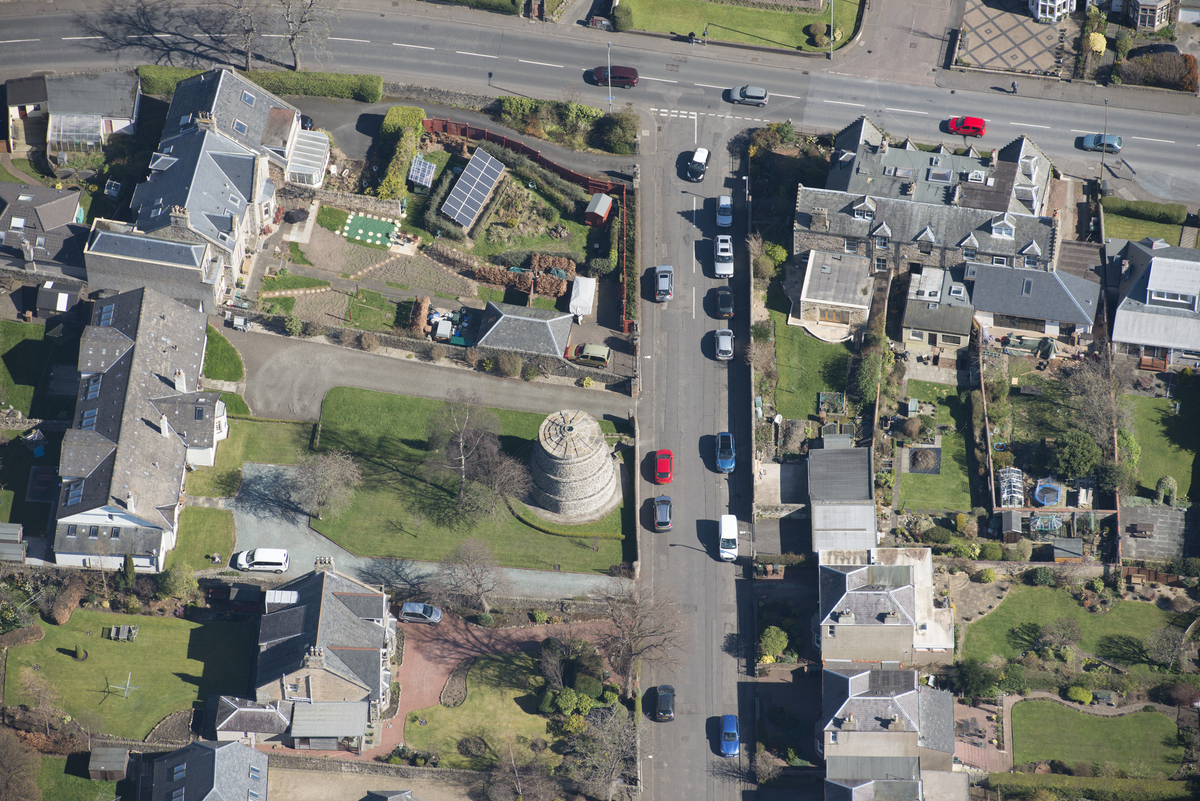 aerial view of a circular stone dovecote with a conical, tiered roof in a residential area surrounded by garden plots, greenhouses, and outbuildings. Two roads run across the image with a few parked cars.