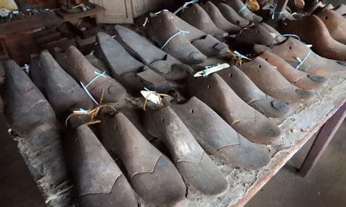 Two rows of old wooden foot models for making and fitting shoes are displayed on a worn table. The models are tagged and there is more shoemaker’s equipment in the background.