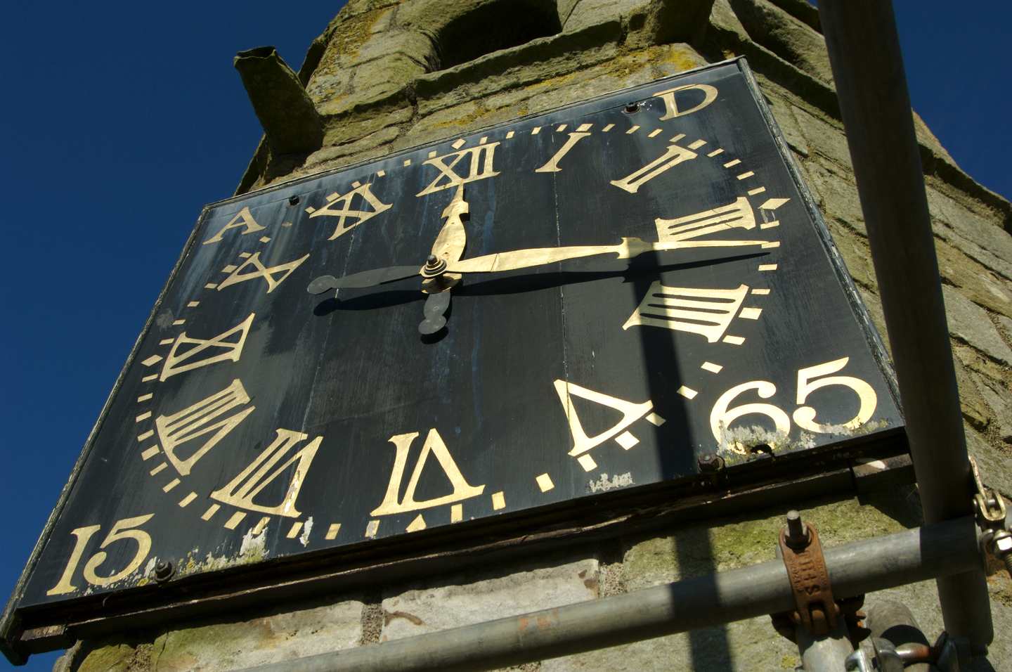 A black clock face with gold Roman numerals and hands, mounted on a weathered stone wall in sunlight.