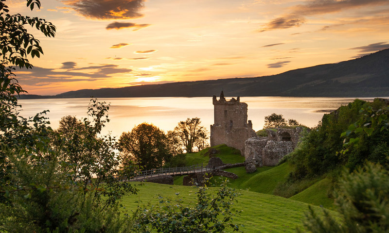 A medieval fortress on the shore of a loch with lush green grass and trees and a view of the loch and hills beyond