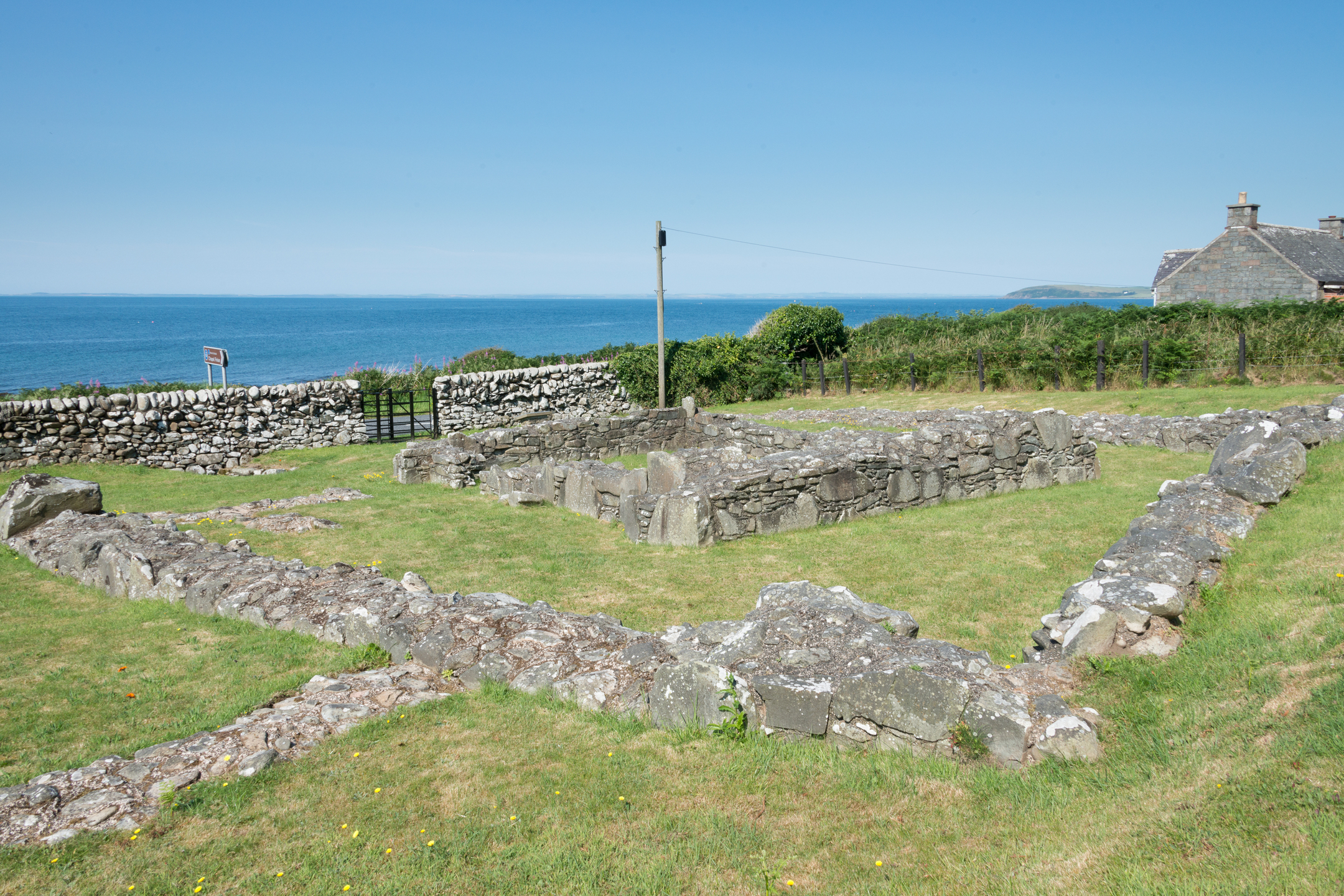 Stone ruins of Chapel Finian forming a rectangular outline on grassy ground, enclosed by a low stone wall. The remains consist of weathered stone walls with uneven surfaces. In the background, the blue sea stretches to the horizon under a clear sky, and a traditional stone building is visible to the right.”