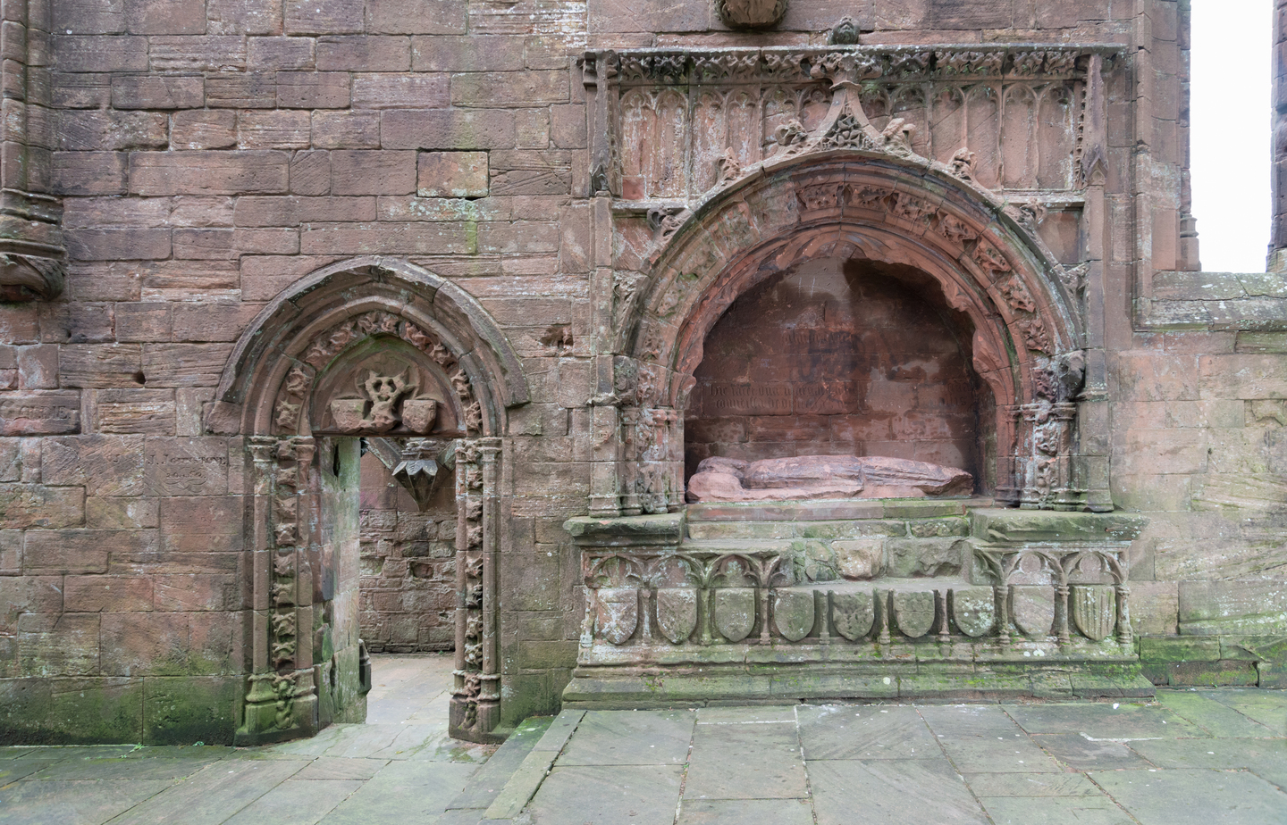 Close-up view of a historic stone wall. You can see a doorway with stone carvings on the left, on the right, you can see a stone effigy of a person laying down. You can see moss.