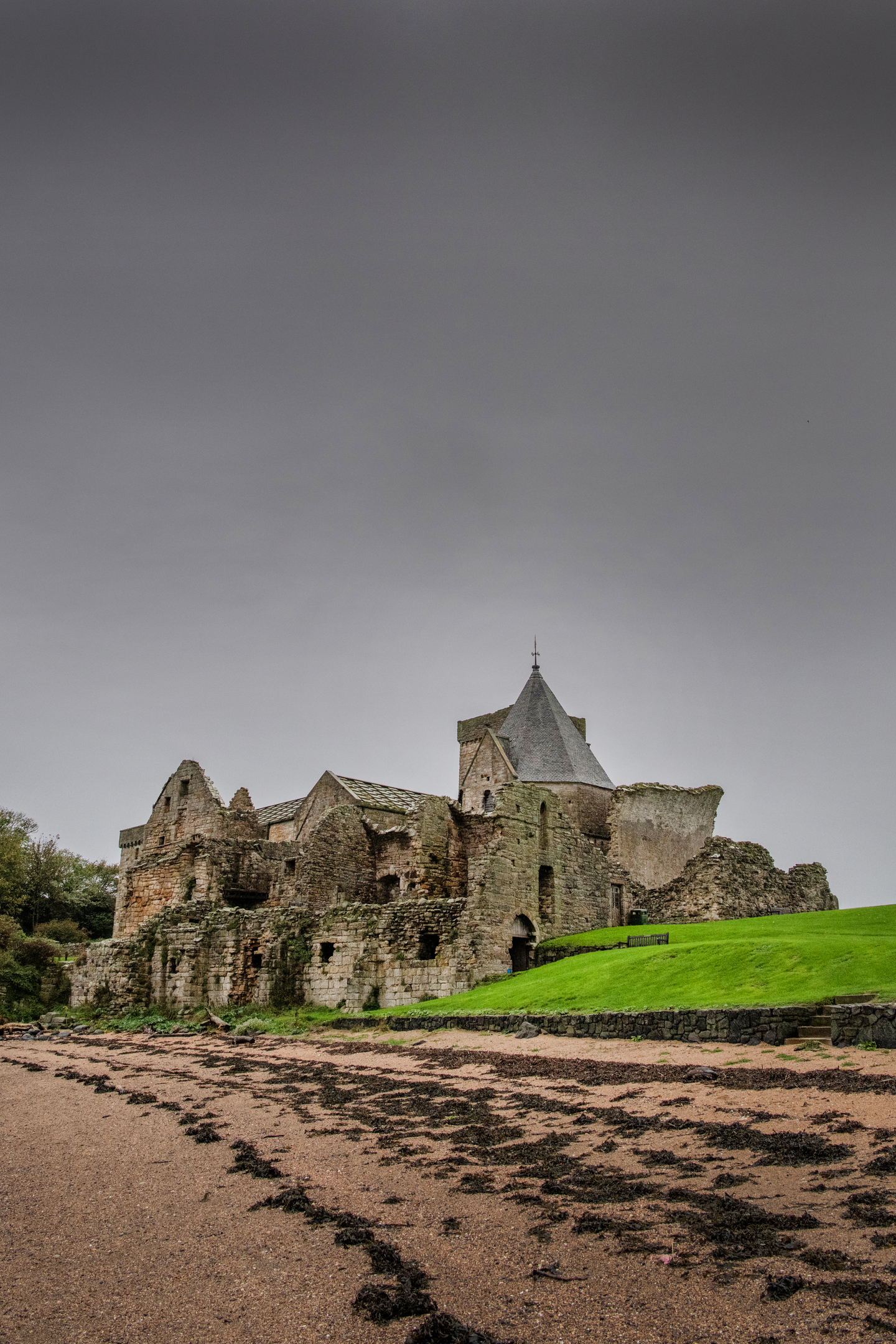 A large, historic stone abbey on a dark, cloudy day beside a sandy beach covered in seaweed
