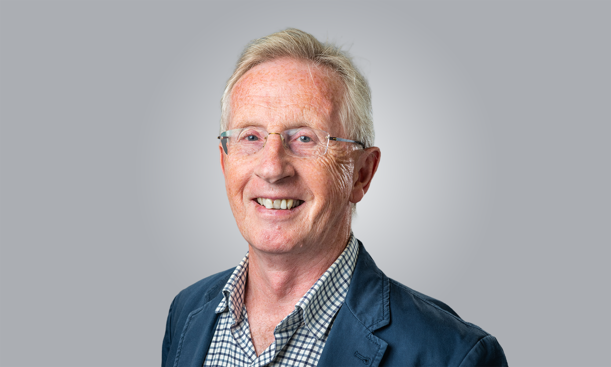 Professional head-and-shoulders studio portrait of a person wearing glasses, a blue blazer, and a checked shirt, facing the camera against a plain light grey background. The image is well lit, with the subject positioned slightly off-centre and looking directly at the camera.