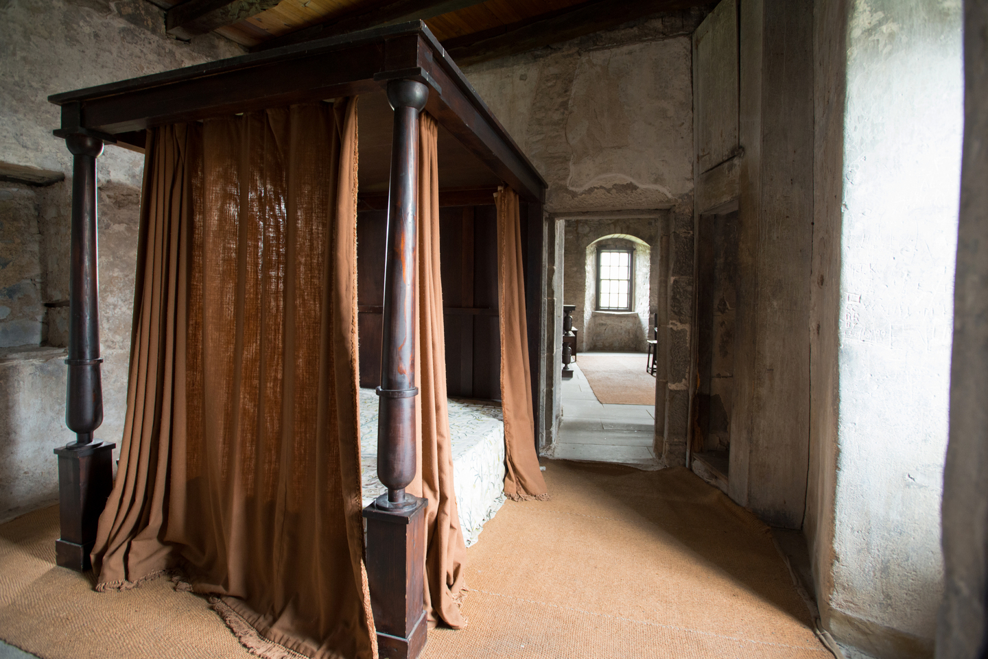 An old room, with pale stone walls. There is a brown carpet. In the room is a historical double bed, with wood beams and brown curtains around the bed.