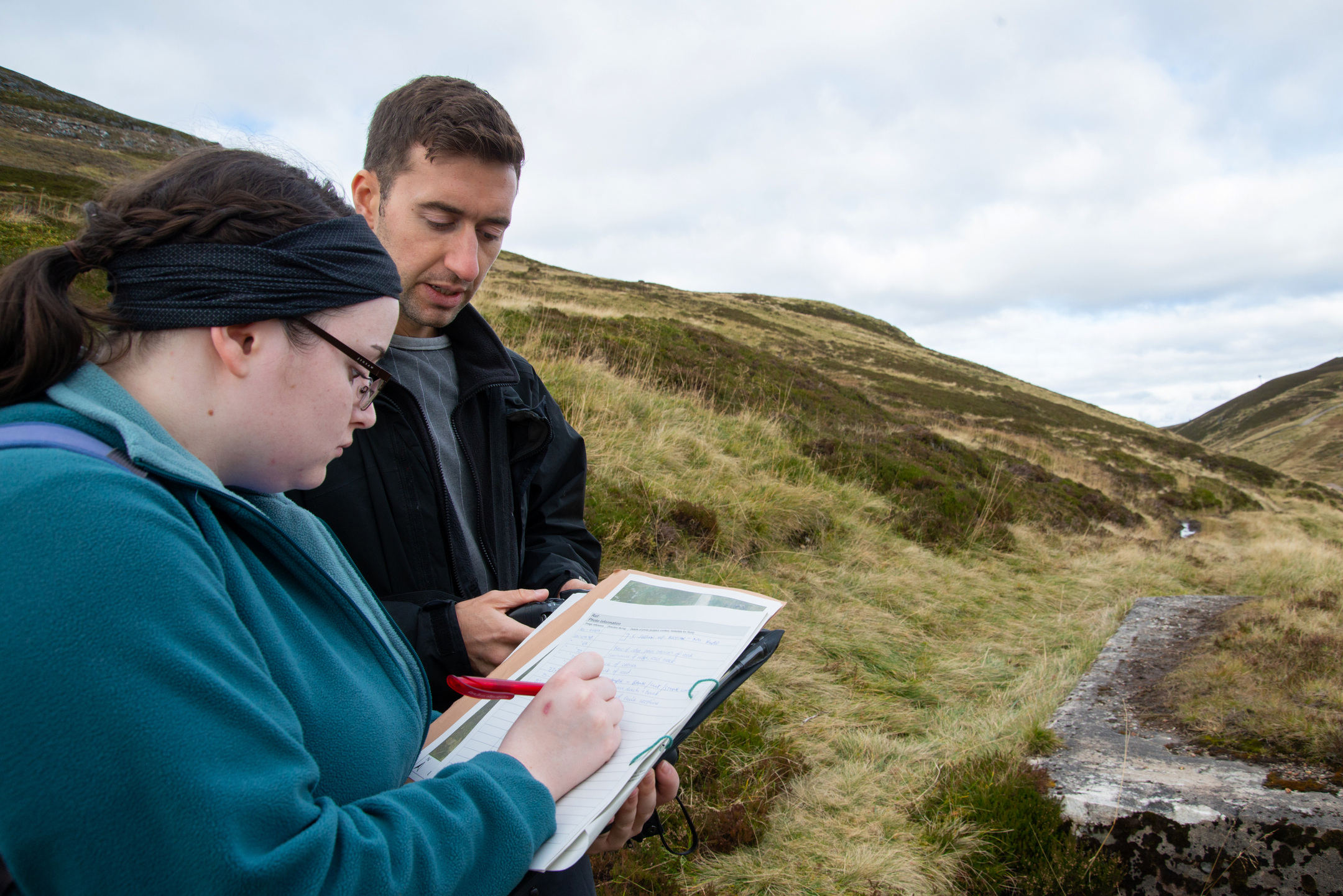 Two people carrying out fieldwork in a grassy upland landscape; one writes on a clipboard while the other looks at a handheld device, with rolling hills and a stone structure nearby under a cloudy sky.