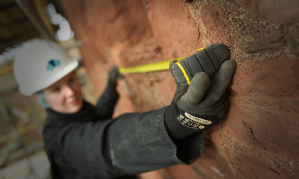 a stonemason in gloves and hard hat holding a tape measure up to an old wall