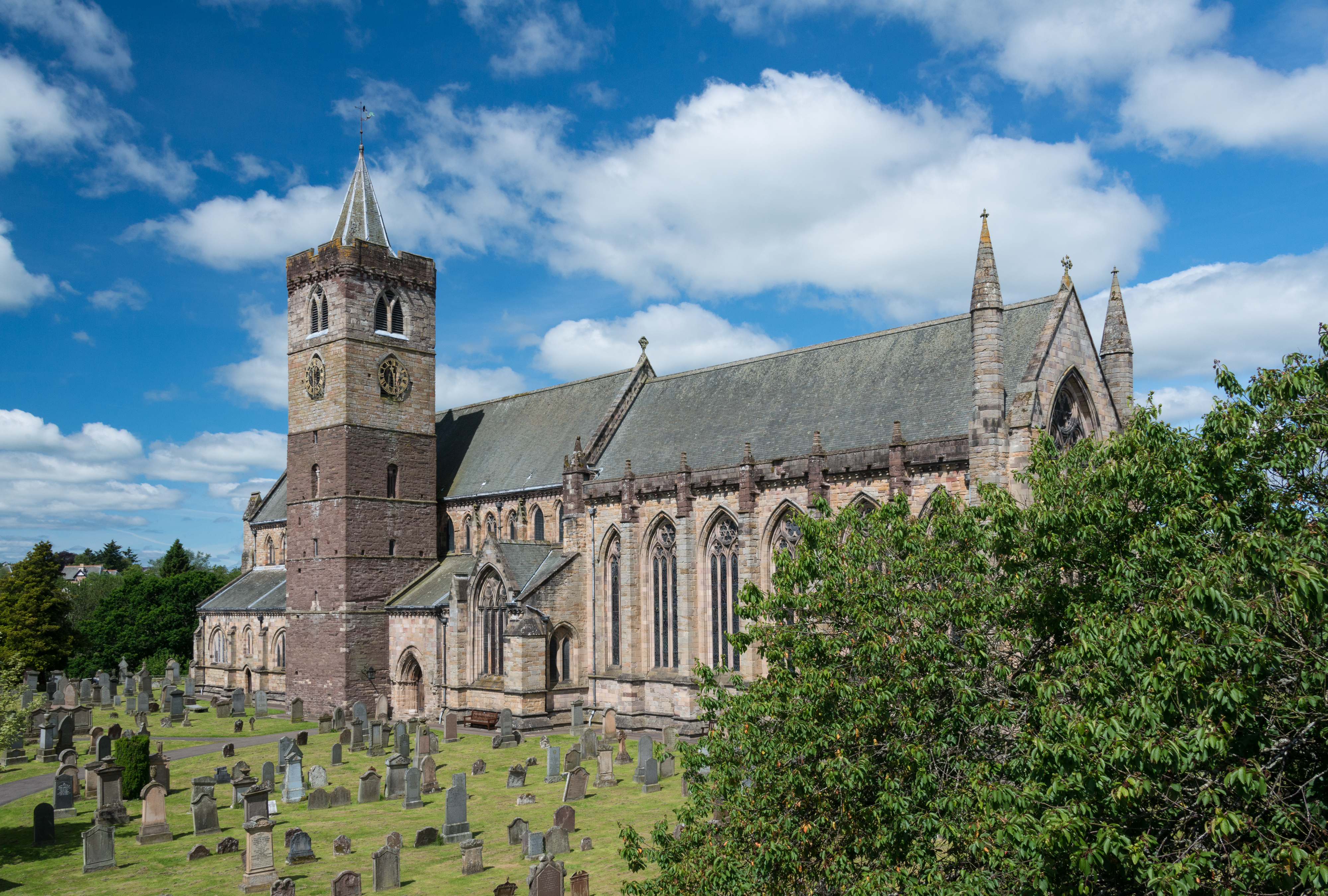 Cathedral with tower and graveyard under a blue sky with clouds. A tree can be seen to the right. 