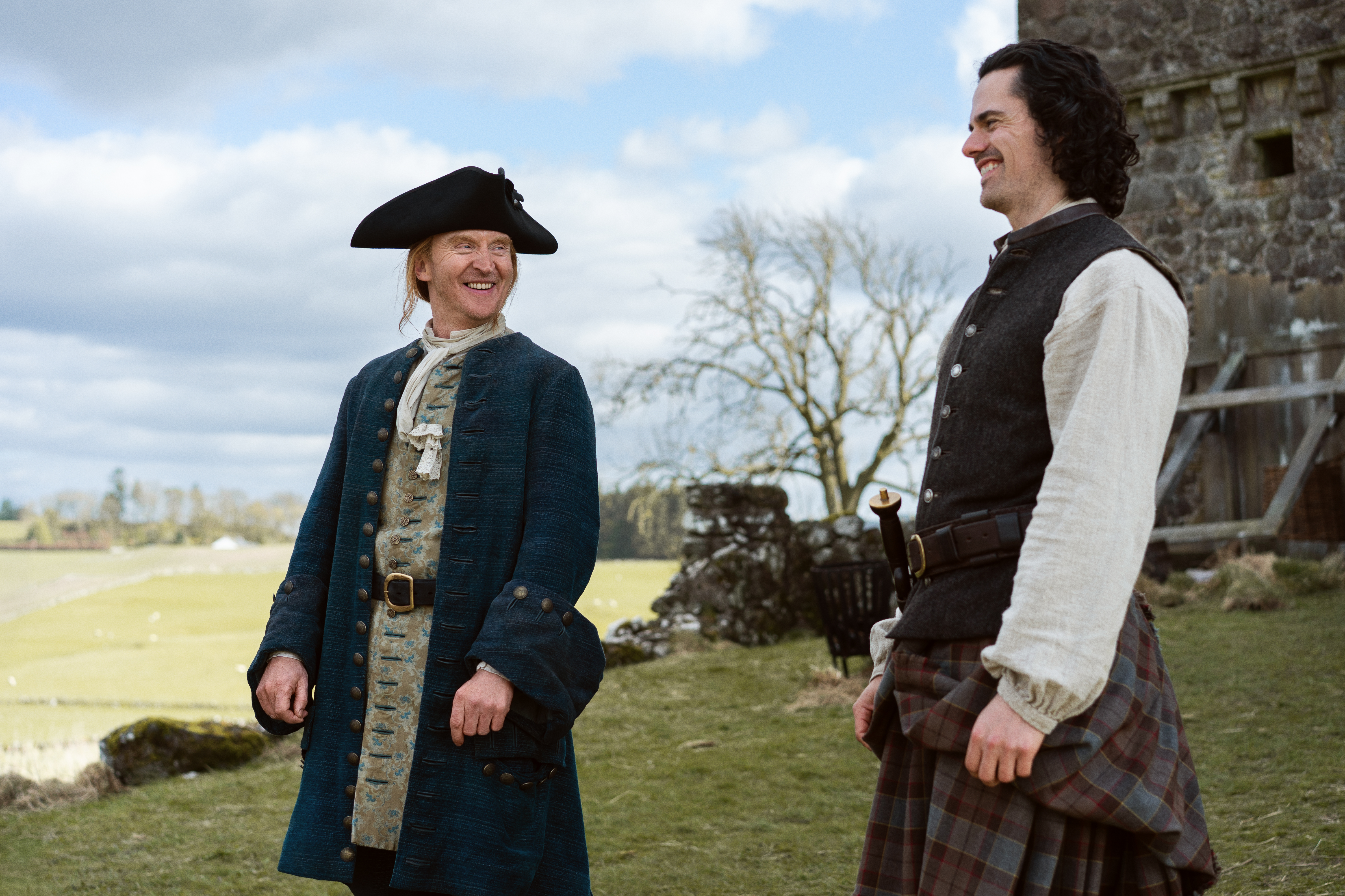 Two actors in period costume pictured outside a ruined castle with fields in the distance.