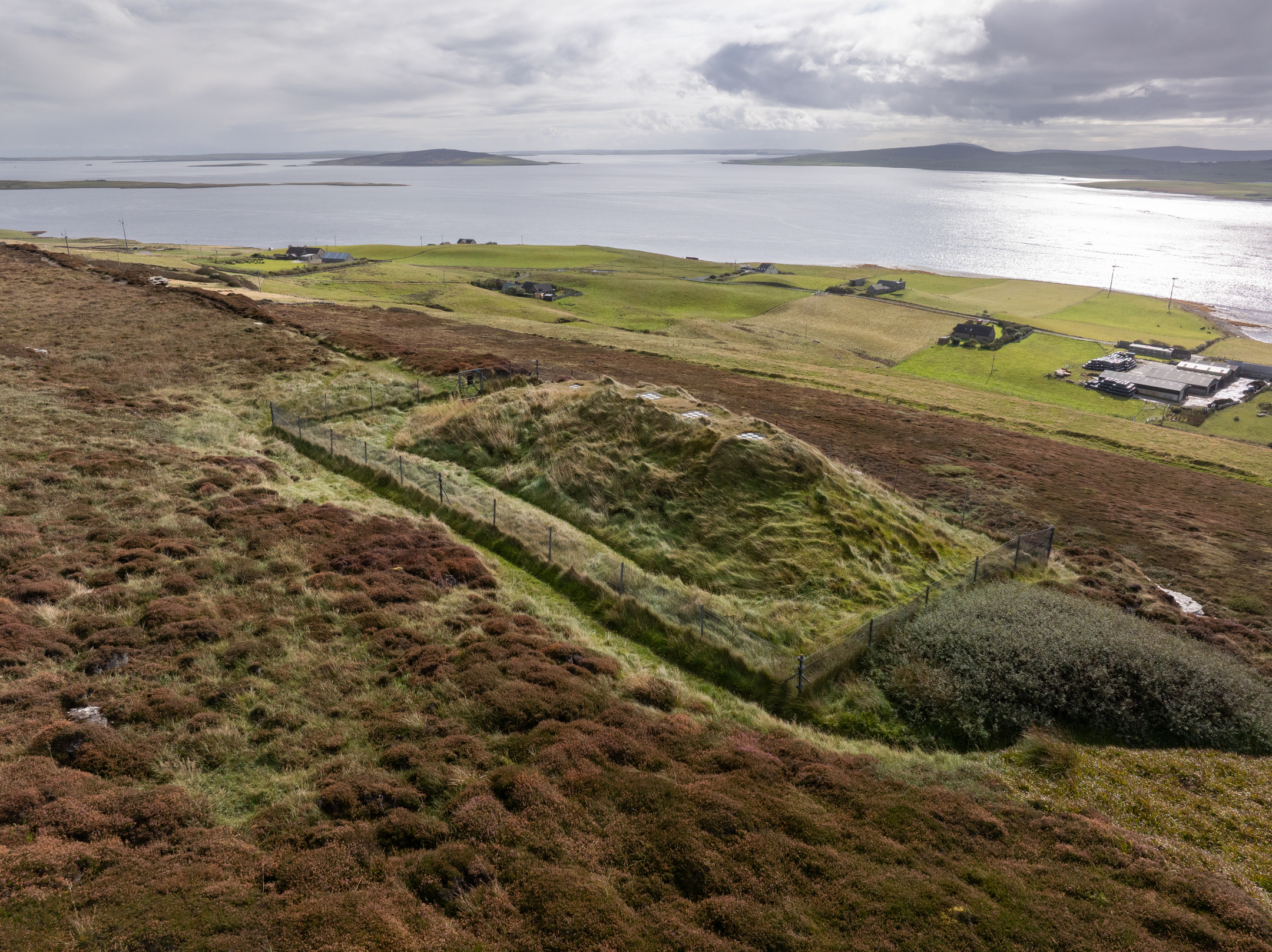 An aerial photo of a grass-covered mount covering a chambered cairn. A panoramic view in the background includes the sea and several small islands.