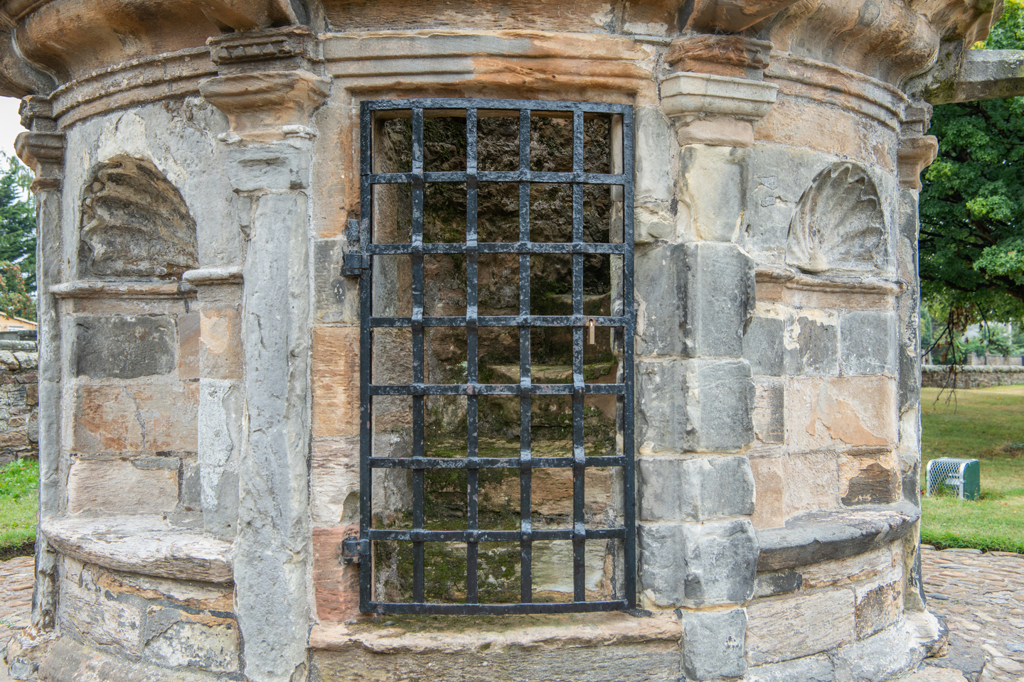 A doorway with a black gate in front of it, with stairs beyond the gate. It is part of a circular stone building. There is greenery in the background. 