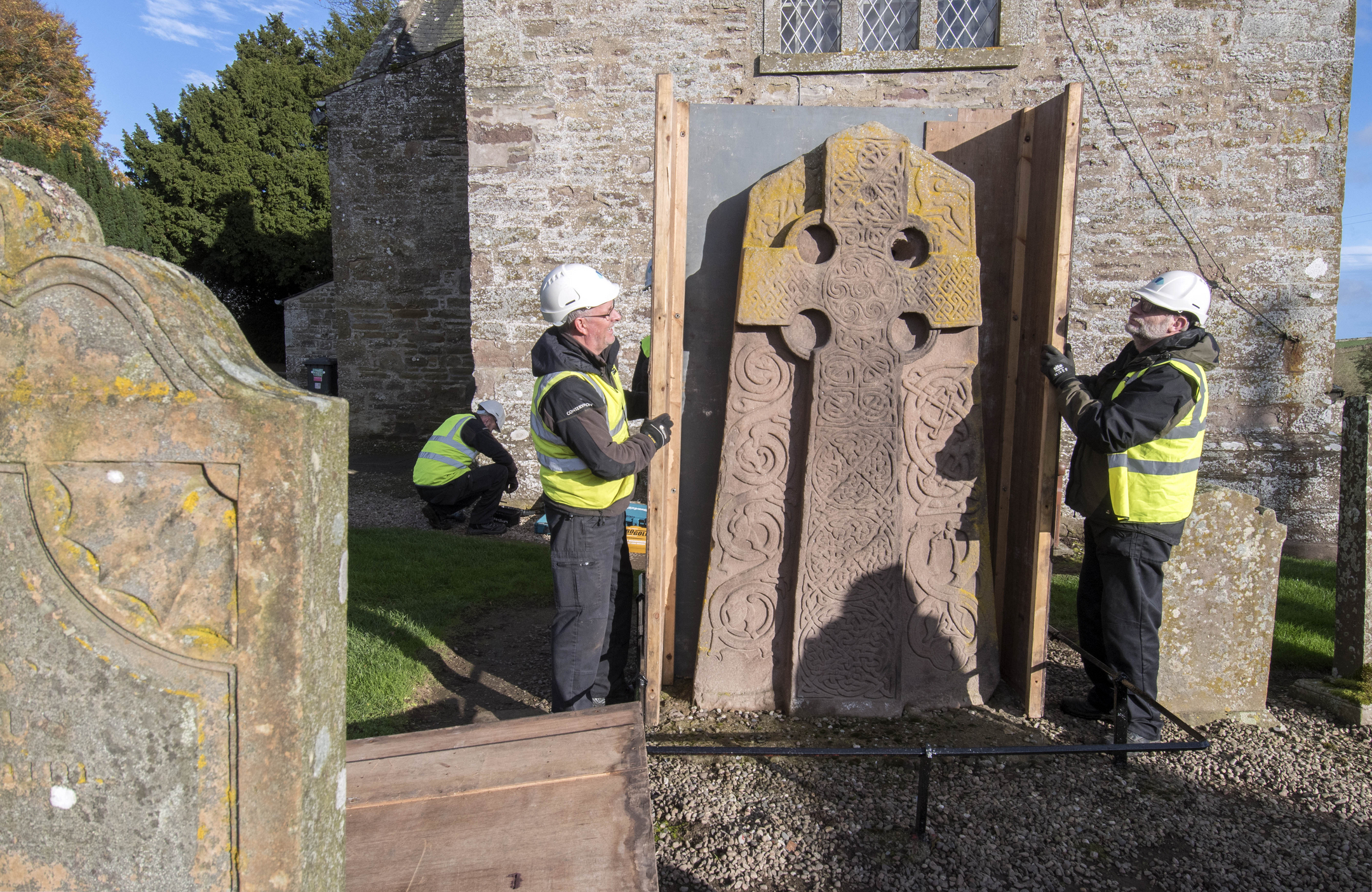 Two workers stand either side of a carved stone in a churchyard. They are holding large wooden panels, ready to erect a protective box around the stone.