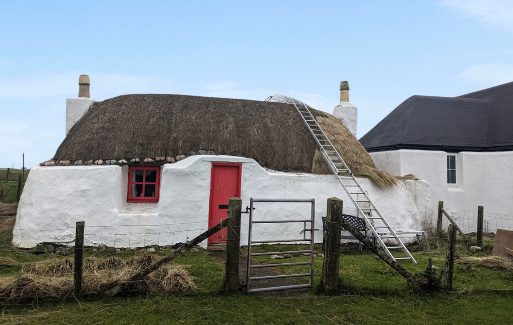 A small stone built, thatched, lime washed cottage with a red front door. A ladder leans on the roof and it looks like the cottage is half way through being re-thatched.