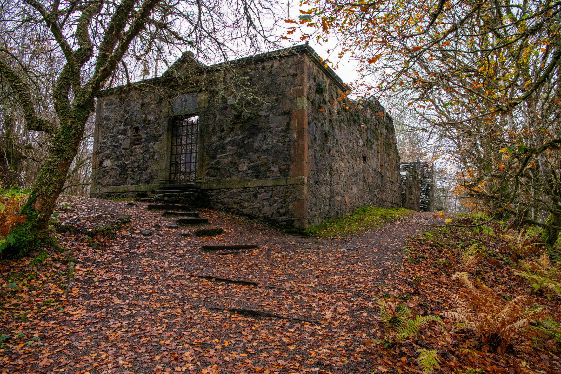 A rectangular stone building ruin in a forest, approached by a leaf‑covered path and surrounded by autumn foliage.
