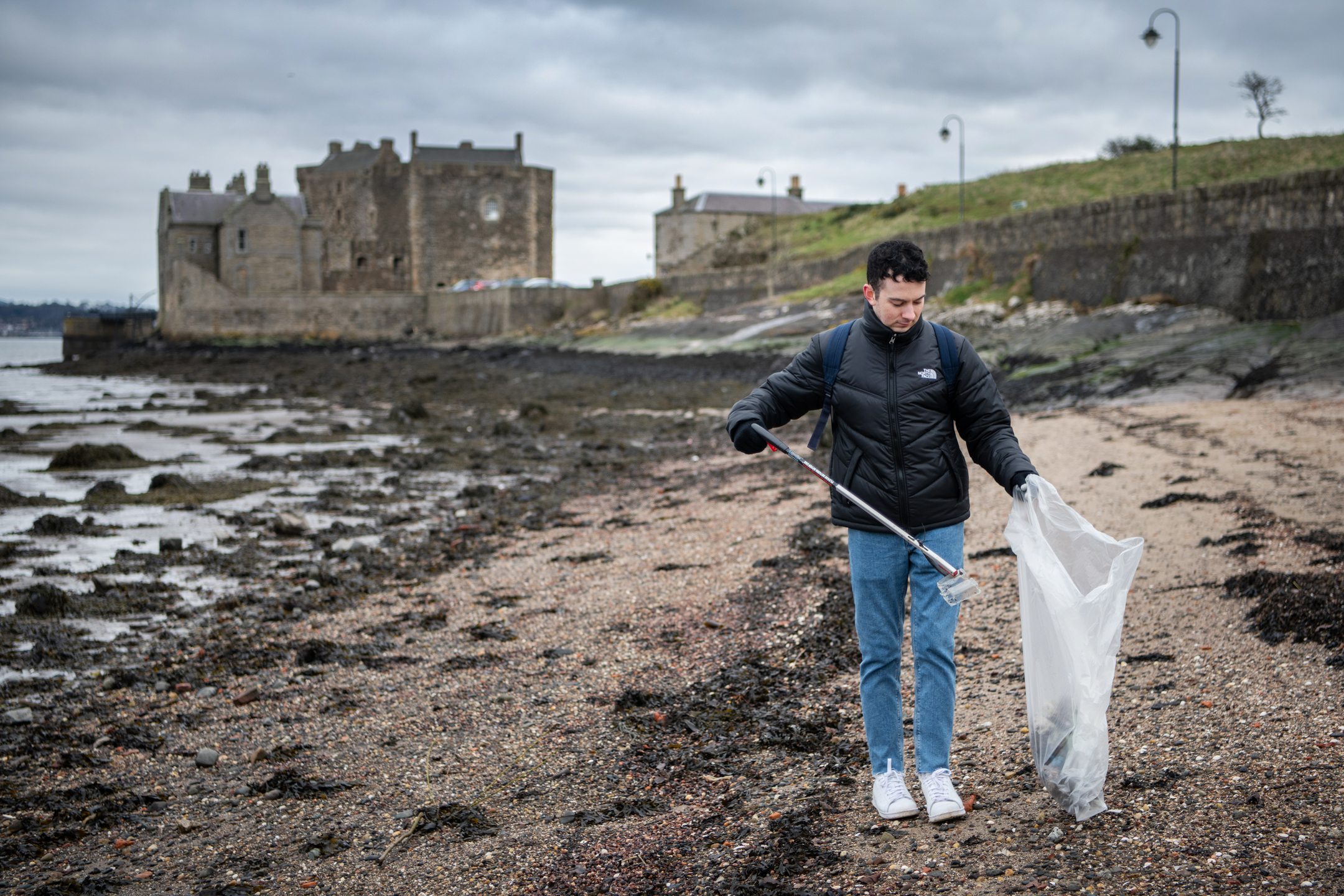 Person uses a litter picker to collect rubbish into a bag on a rocky beach, with a large stone building and cloudy sky in the background.