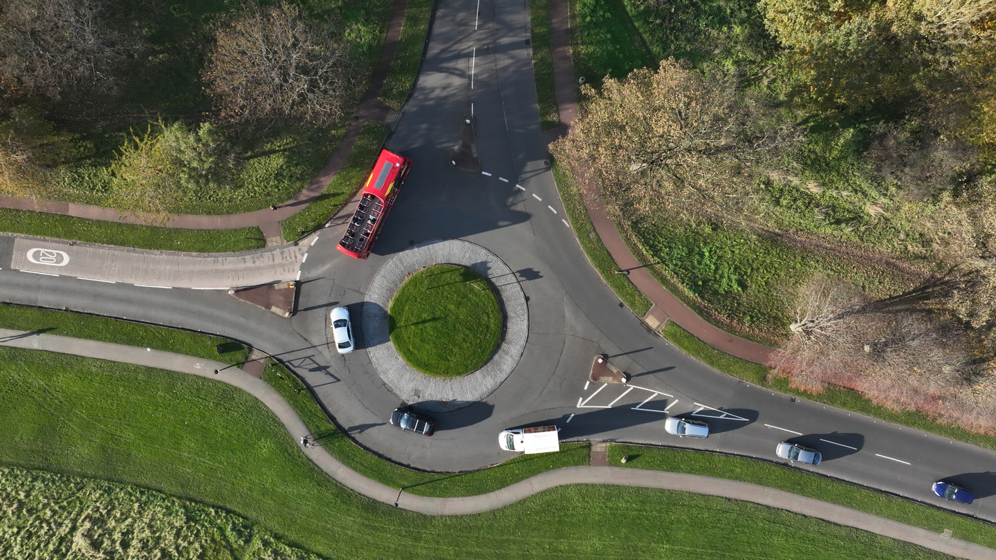 Aerial view of a small roundabout in Holyrood Park, showing several cars and a red open‑top tour bus travelling around it. The roundabout is surrounded by curved roads, green grass, and tree‑lined paths.