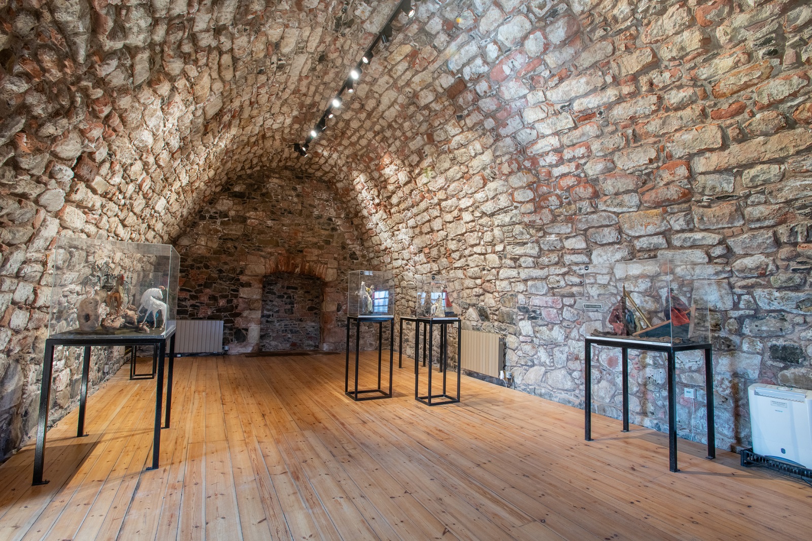 Interior of a vaulted stone room with wooden floor and display cases showing small artefacts.