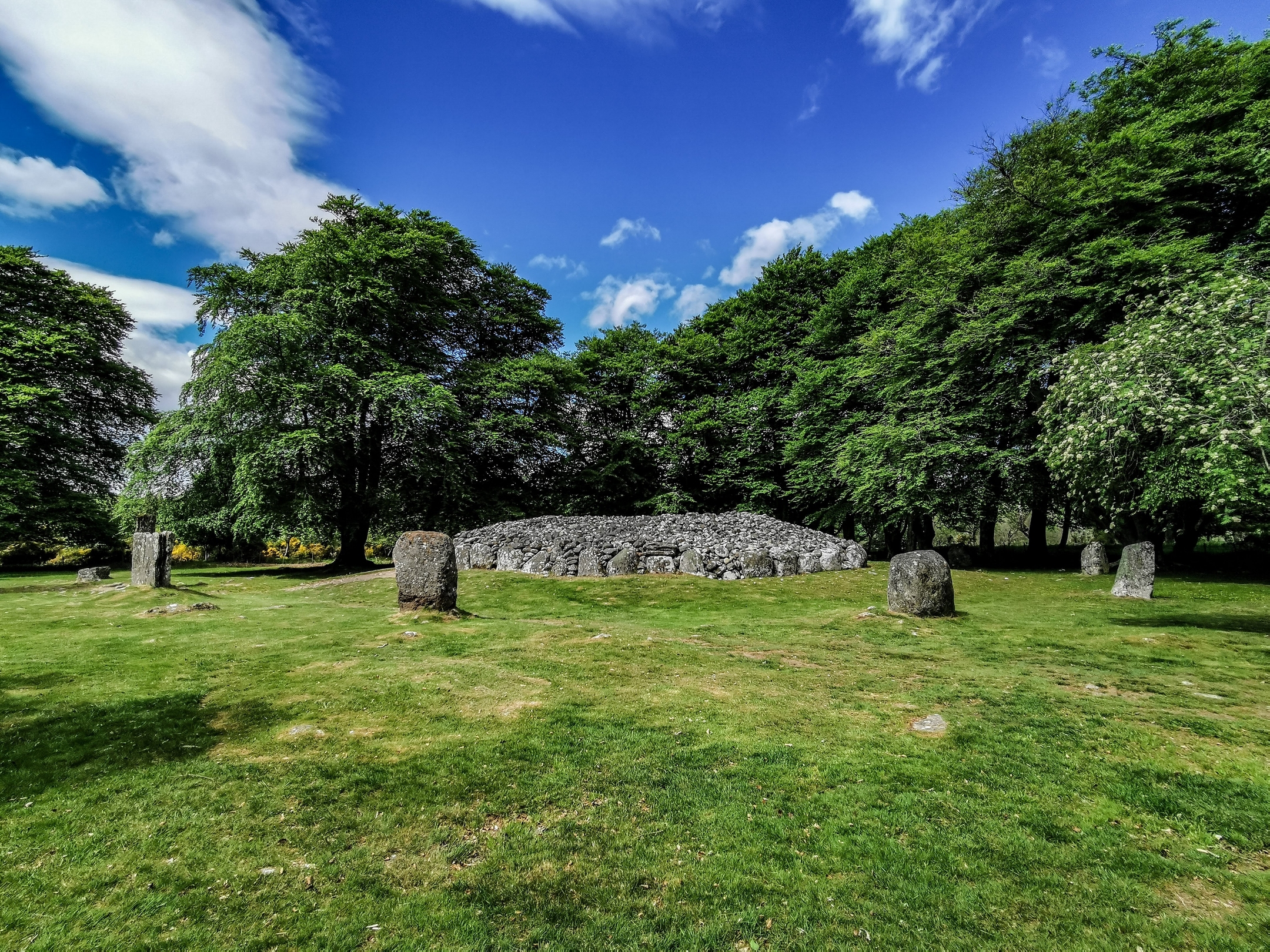 Blue sky, green trees and grass, and a cairn, centre, with standing stones around it