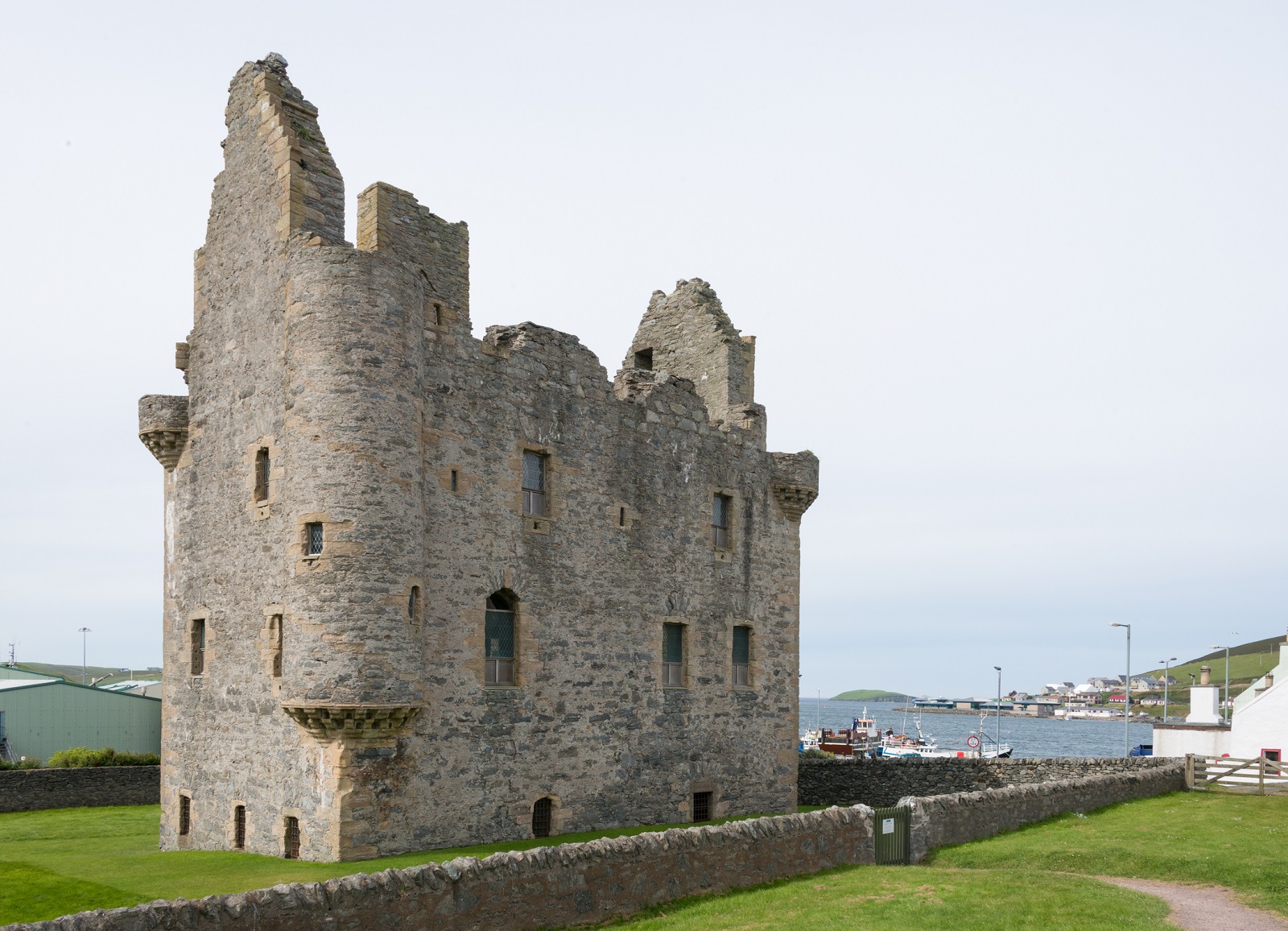 Side view of Scalloway Castle’s tall, roofless stone walls and corner turrets, with a low stone boundary wall in front and the harbour visible in the background