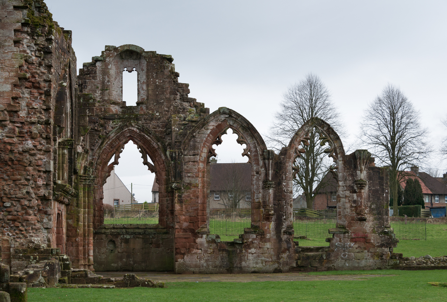 Ruins of part of a historic stone building, you can see the remains of tall arched windows and walkways, sat on a grassy landscape. there are trees in the background.