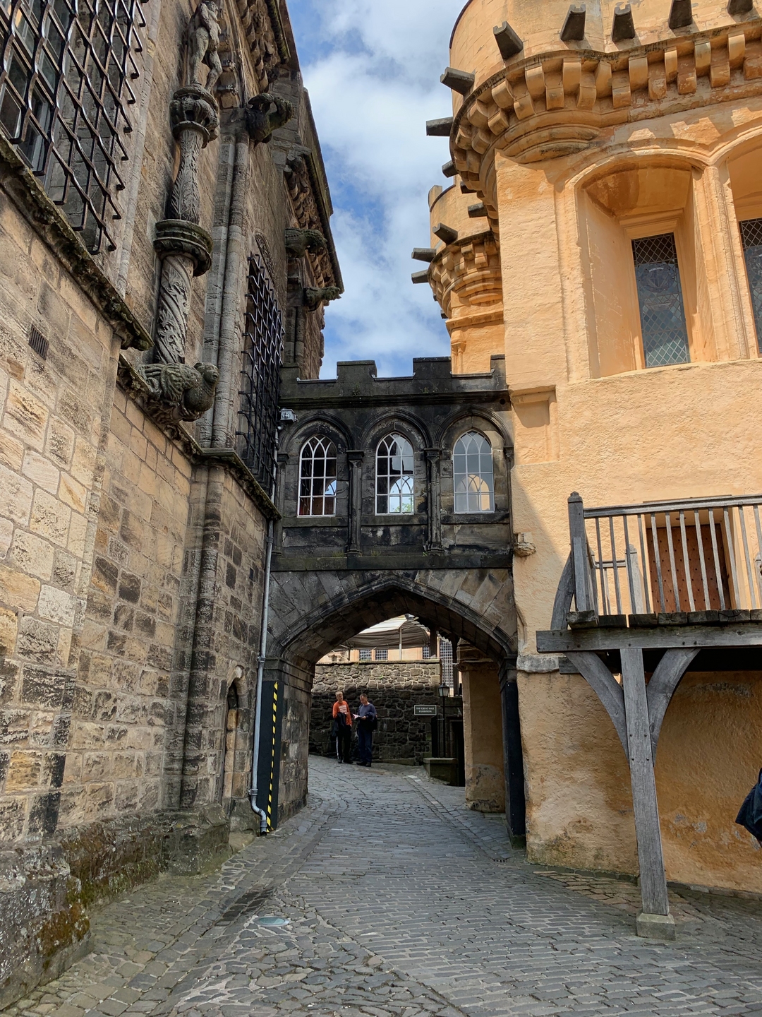 A walk way between two buildings, with a connecting bridge between the two with an arch in the middle. The building on the right is orange, and while not modern, appears more recent than the building on the left. This building, as well as the connecting bridge, is made of stone which has turned black in areas. 