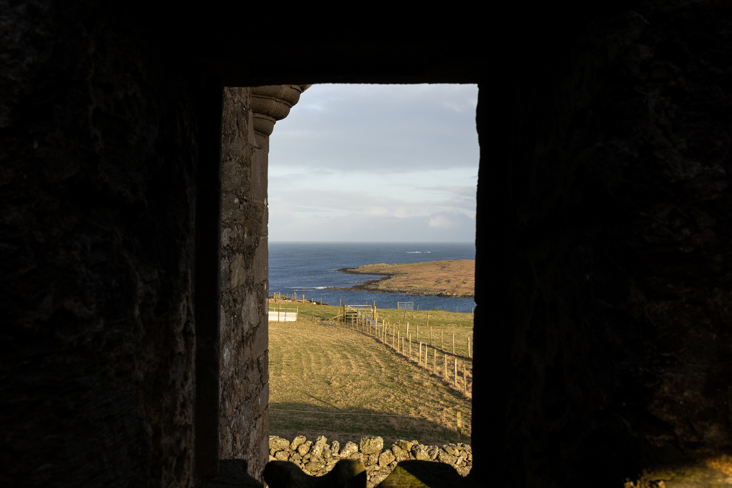 View through a stone-framed window at Muness Castle, looking out onto a grassy field that slopes toward the coastline. 