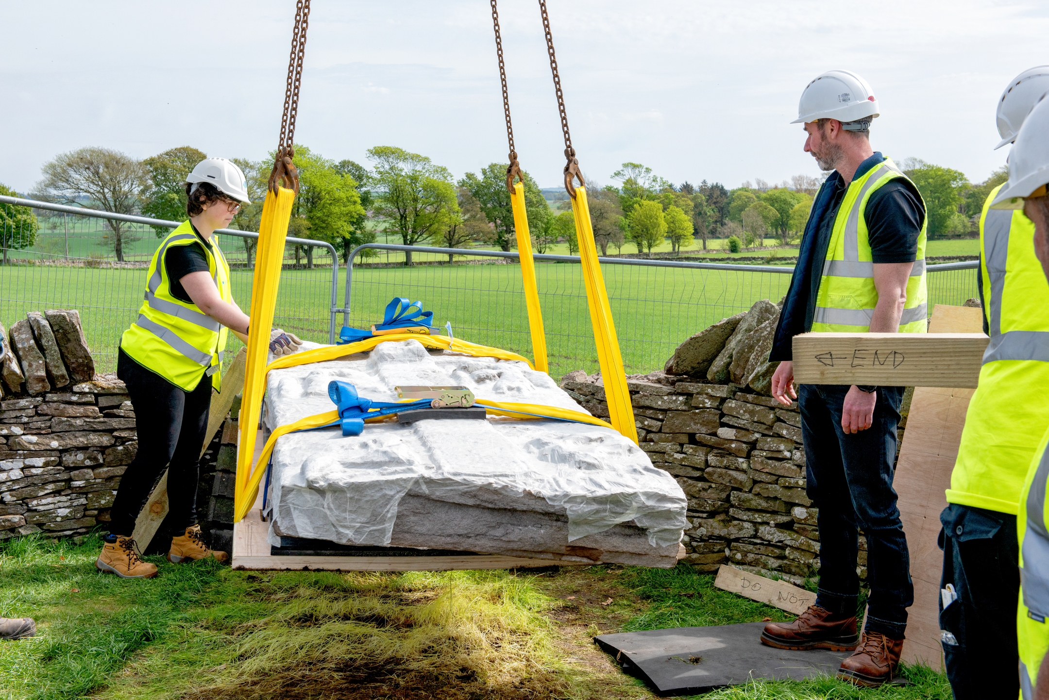 Workers in hi-vis and hard hats stand by as a carved stone is carefully lifted using a crane.