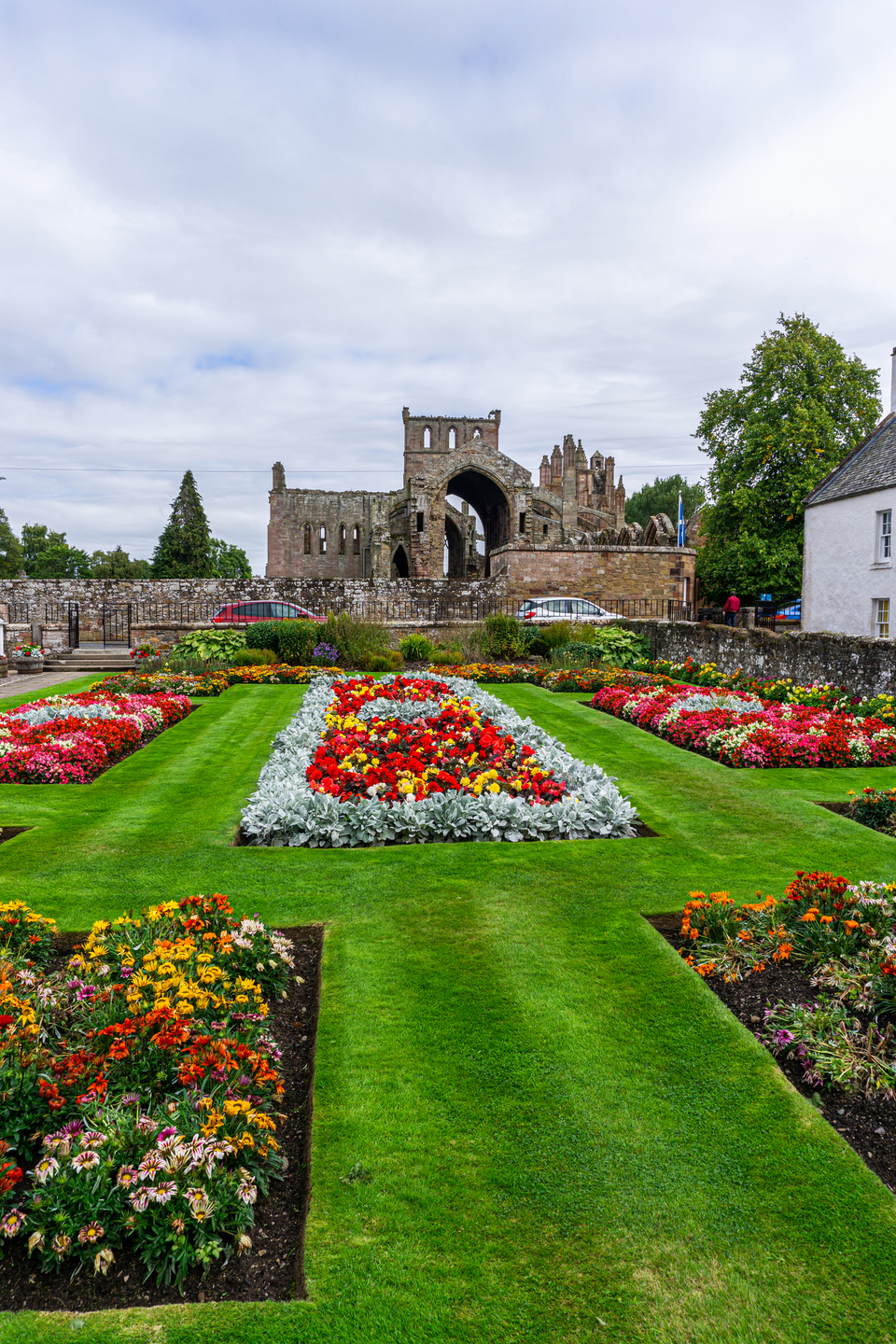 A tidy garden with squares of colourful flower patches. In the background is a tall historical building with large arches. 