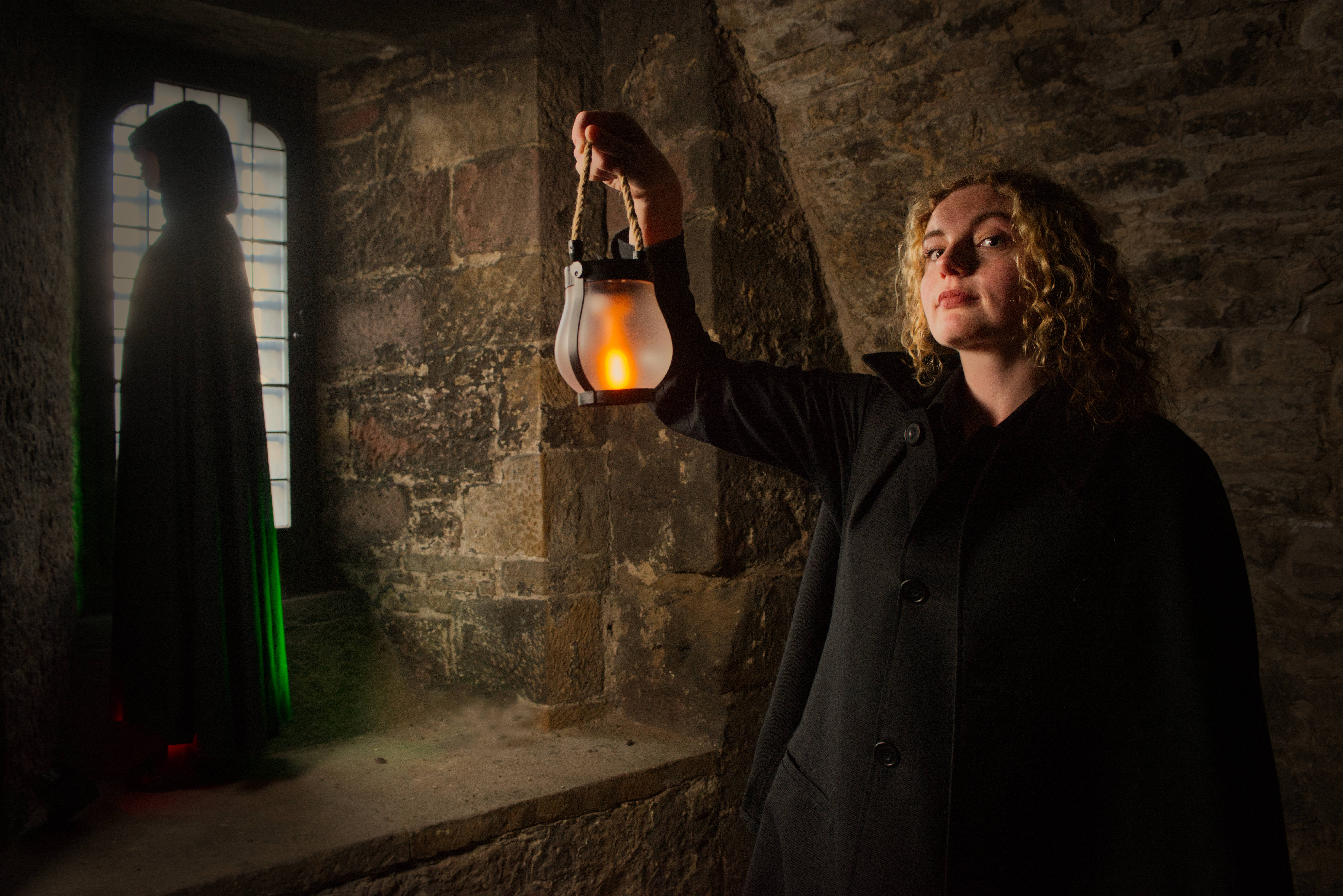 Woman holding a lantern in an old stone room. A cloaked figure can be seen beside her.