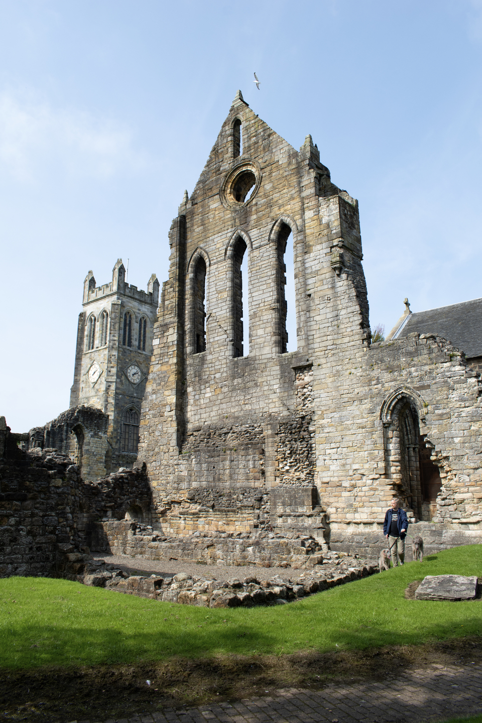 Ruined wall of a stone church with three narrow arched window openings and a circular opening above them. To the left is a square clock tower, part of a newer building still in use. At ground level are low stone foundations.