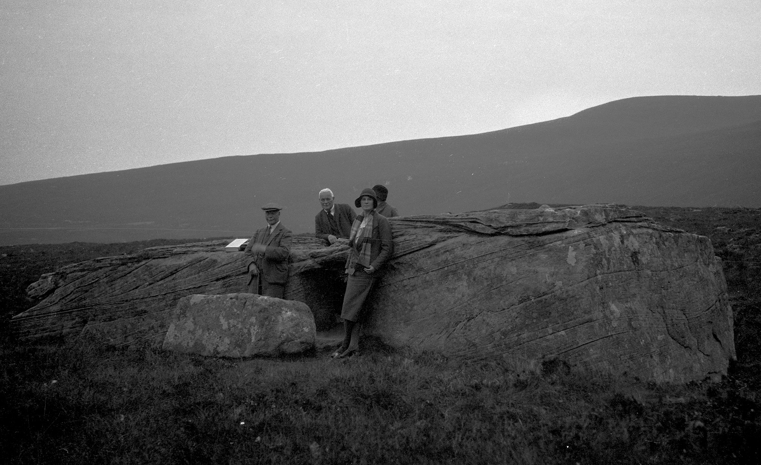 A black‑and‑white 1920s photograph of four people standing beside the Dwarfie Stane, a large rectangular boulder with a carved entrance. The group is dressed in period outdoor clothing, and low, rounded hills rise in the background under an overcast sky.