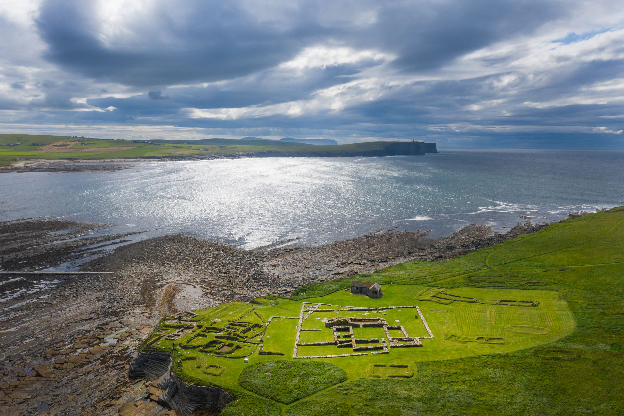 A wide drone view of the Brough of Birsay ruins on vivid green grass, with the sea shimmering under dramatic cloudy light and distant cliffs along the coast.