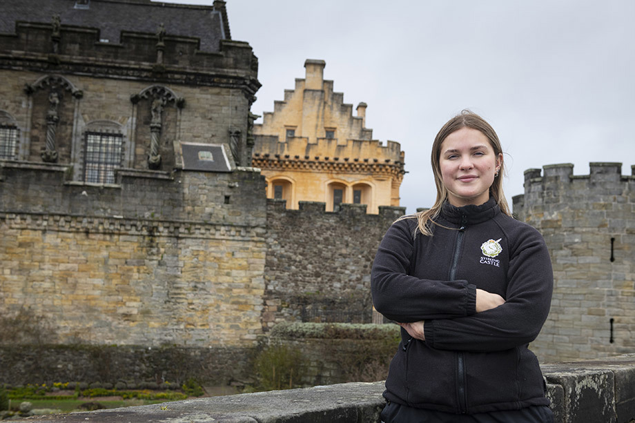 A young woman in a black zip up fleece with a 'Stirling Castle' logo on the chest stands arms folded in front of a castle.