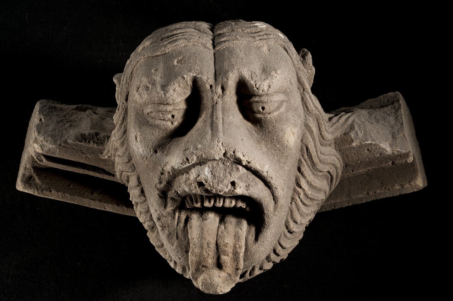 An unusual carved stone head on a black background. It resembles an animal with human features, with a mane around the face. It is looking down with its tongue out.