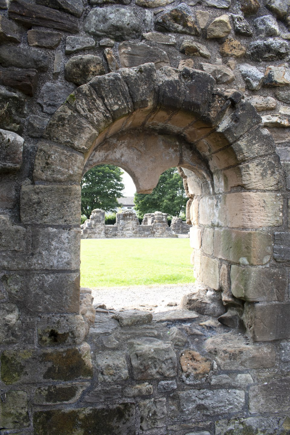 View through a small arched stone window set in rough, weathered stonework. The view looks onto a grassy area with low stone walls and trees behind them.